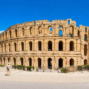 esim data trip for Tunisia to visit the  ancient Roman Colosseum in El Djem, Tunisia, under a clear blue sky, with a small tourist walking in the foreground plaza.