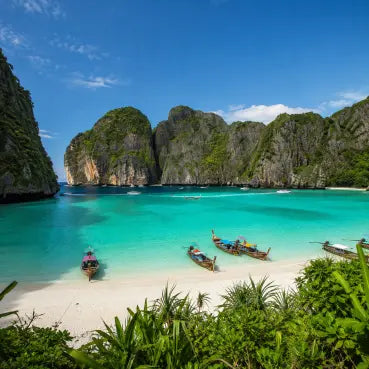 eSIM Data trip to Thailand, the picture with scenic tropical beach with turquoise water, white sand, tall green cliffs, and several longtail boats on the shore under a blue sky.